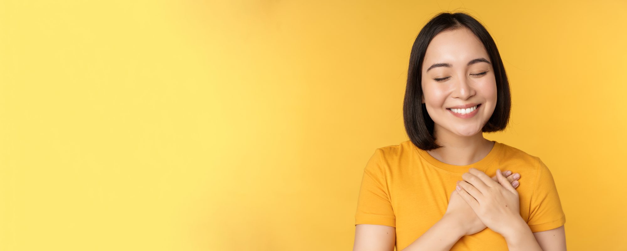 Elyxa Australian Organic Whole-food Supplements Beautiful Asian Woman, Smiling With Tenderness And Care, Holding Hands On Heart, Standing In Tshirt Over Yellow Background